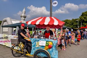 Familien.Spiele.Fest in Köln | Im Vordergrund ein Fahrrad-Eiswagen. Ein Mann sitzt auf dem Fahrrad und schaut freundlich in die Kamera. Eine Frau steht hinter dem Wagen und bietet das Eis an.